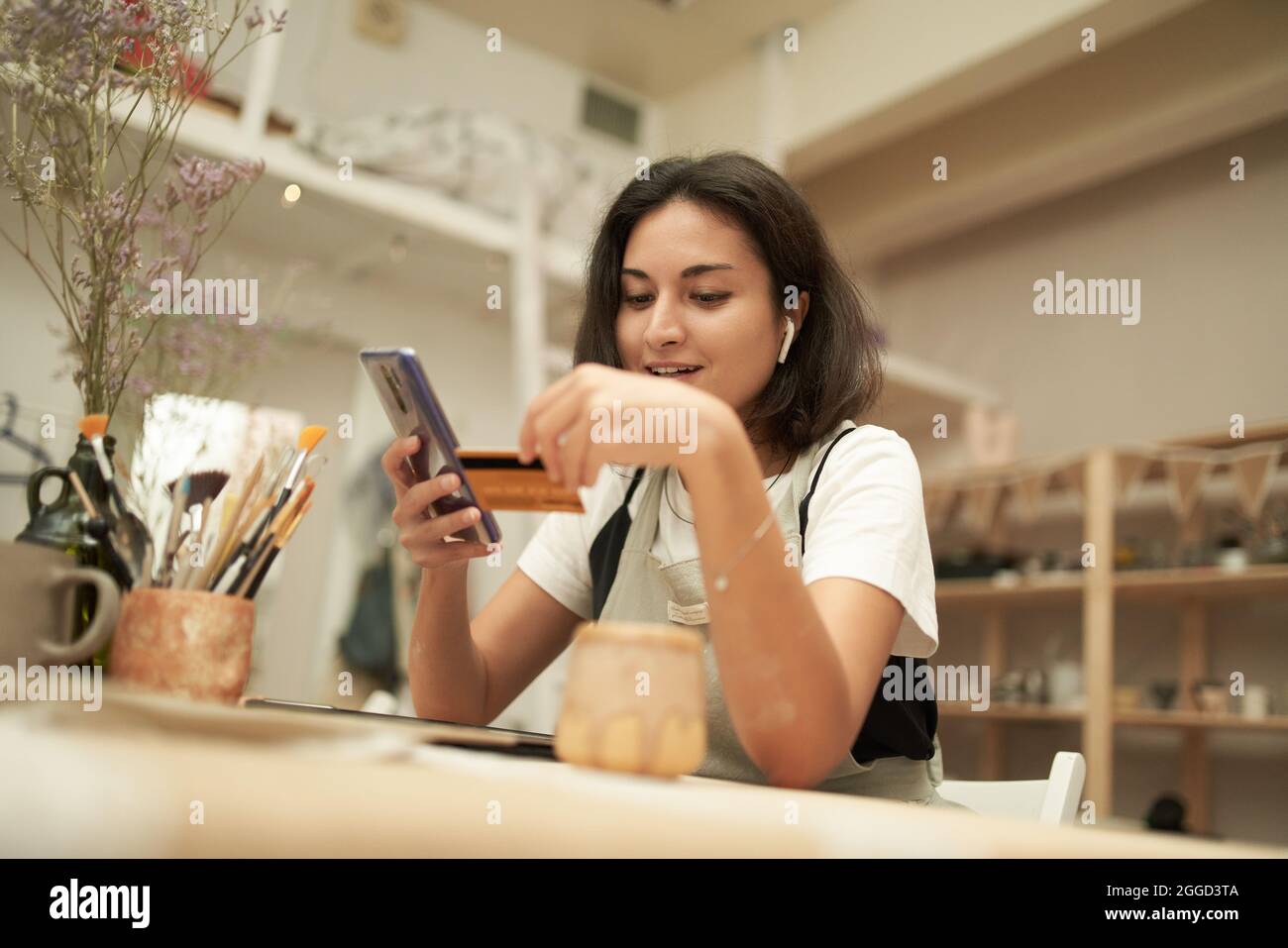 Artigiano femminile che inserisce le credenziali della carta di credito nel telefono cellulare mentre paga per l'acquisto in linea durante il lavoro in ceramica professionale Foto Stock