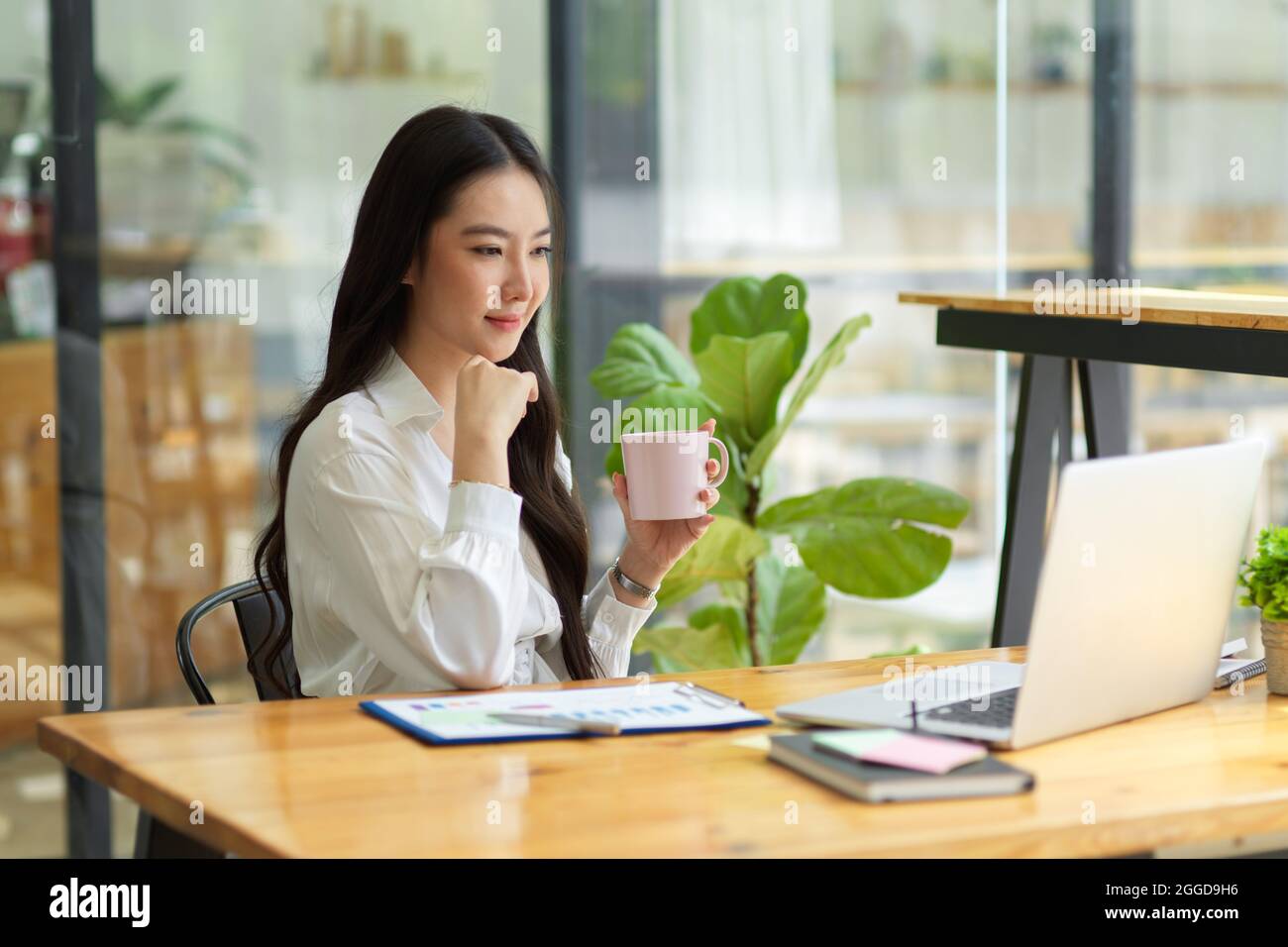 Donna d'affari intelligente che lavora sul laptop mentre beve il caffè nel suo ufficio, attraente auditor finanziario femminile che guarda sul laptop, documenti finanziari su Foto Stock