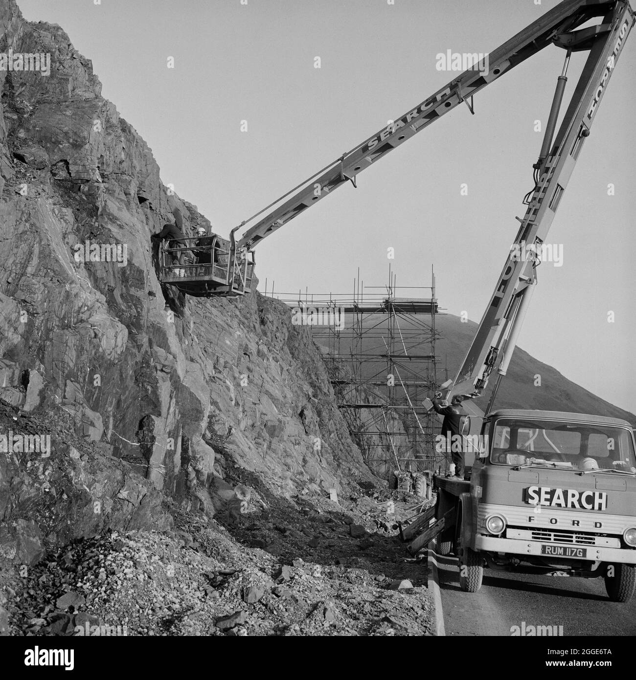 Una vista della costruzione dell'autostrada M6 attraverso la Gola di Lune, che mostra due uomini in una piattaforma di sollevamento idraulica montata su camion, che lavora contro una parete di roccia a sud di Low Borrowbridge. I lavori sulla sezione Lune Gorge dell'autostrada M6 tra Killington e Tebay (Junction 37 - Junction 38) sono stati eseguiti da John Laing Construction Ltd. I lavori sono iniziati nell'ottobre 1967 e l'autostrada è stata aperta al traffico nell'ottobre 1970. Su questo tratto di autostrada, le scogliere di roccia sono state perforate, sabbiate e poi imbullonate per la stabilità. Foto Stock