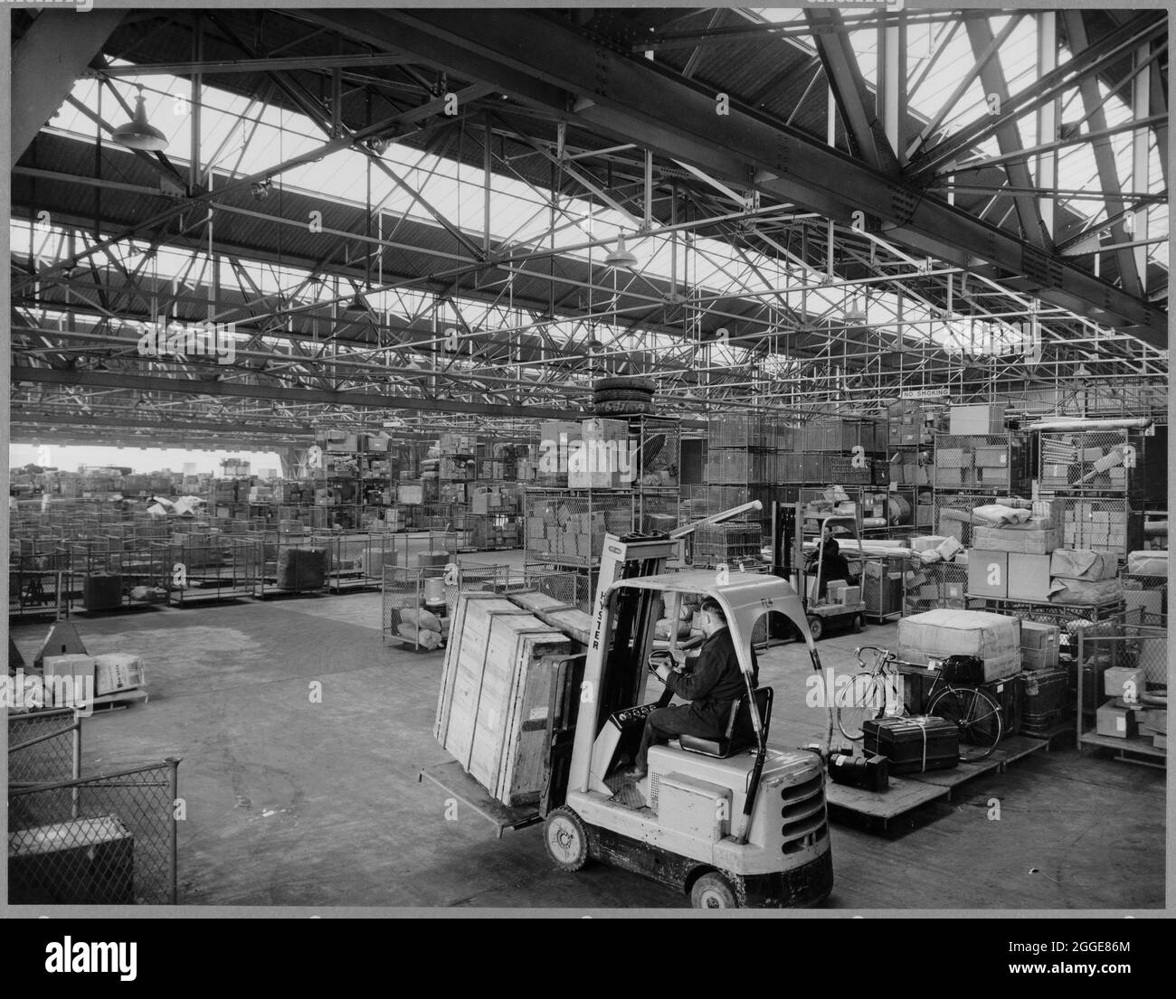 Guardando attraverso l'interno del capannone di trasporto e pacchi presso il British Road Services Depot, mostra un conducente di carrelli elevatori che sposta le casse in primo piano. Il capannone di trasporto e pacchi fu costruito da Laing alla fine degli anni '50 per British Road Services (in seguito National Freight Corporation). Aveva un'ampia area del pavimento senza ostacoli resa possibile dall'uso di massicce travi in traliccio che sostengono la struttura. Foto Stock