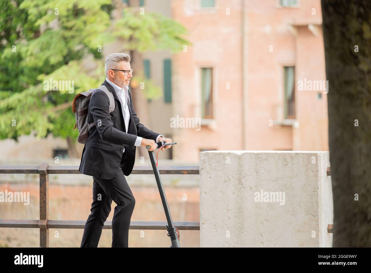 Vista laterale per tutta la lunghezza del giovane uomo d'affari in una tuta che guida uno scooter elettrico mentre si viaggia per lavoro in città. Concetto di trasporto ecologico Foto Stock