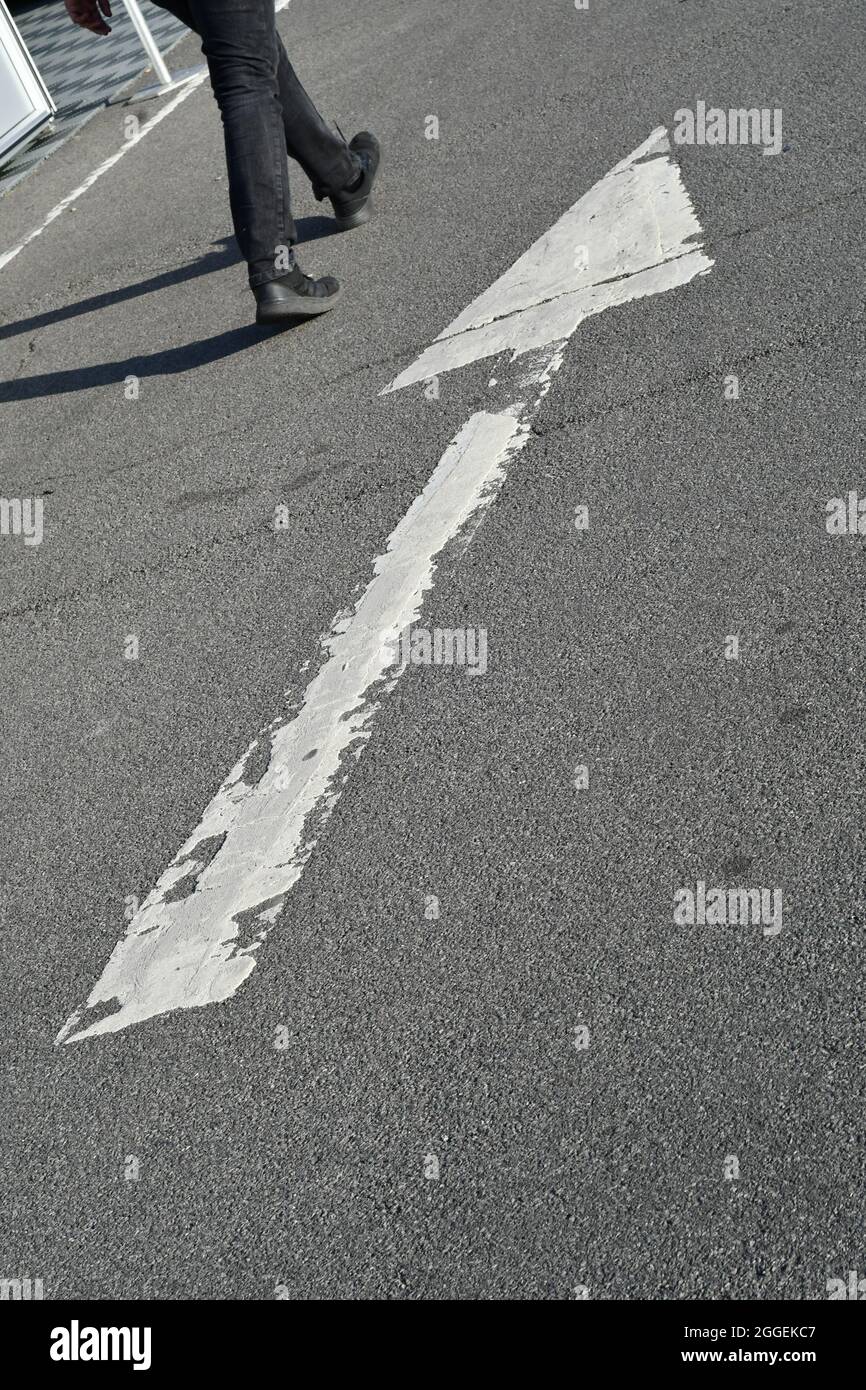 Un uomo che cammina lungo una strada contrassegnata da una grande freccia bianca usurata Foto Stock
