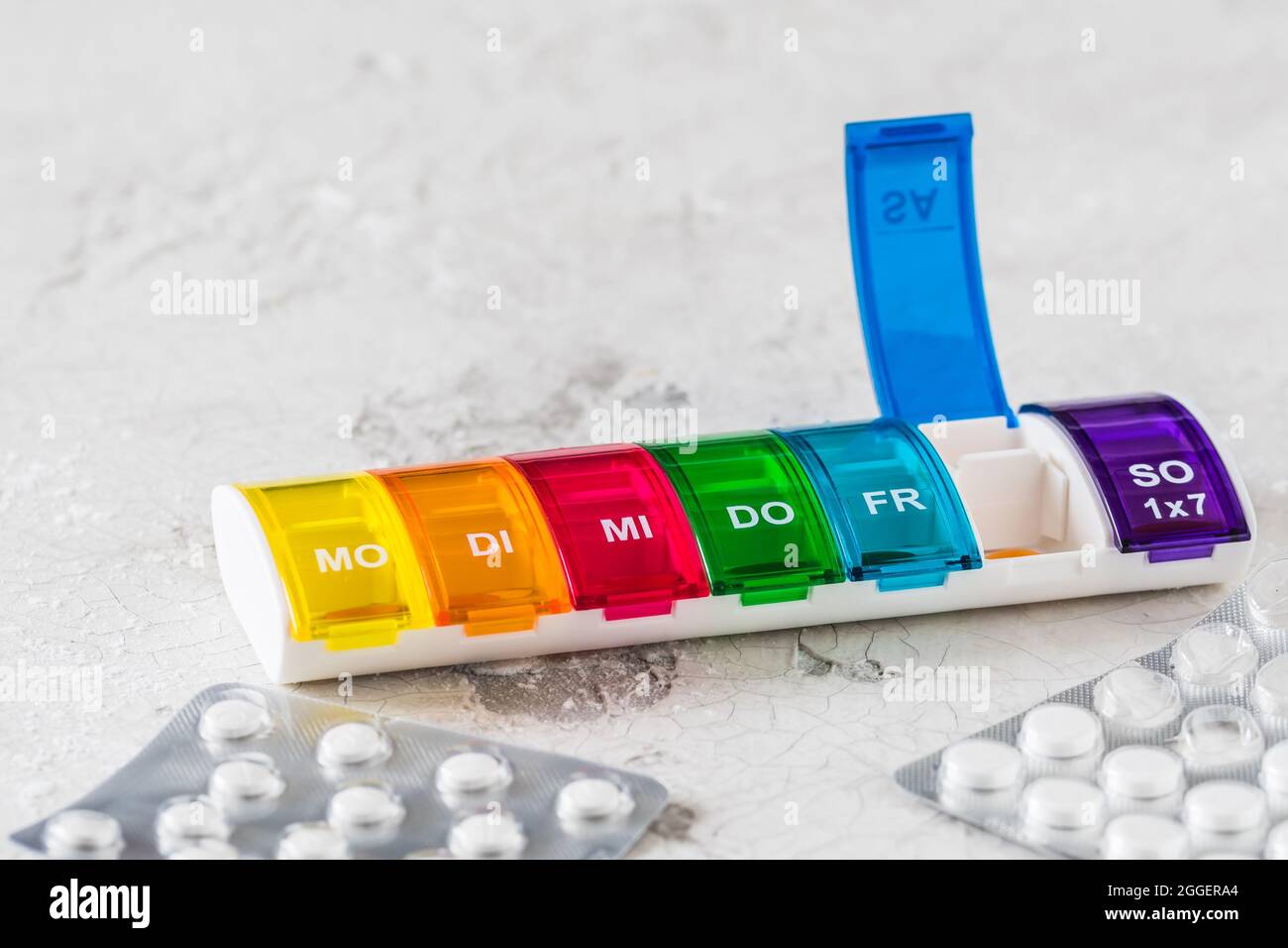 Scatola di pillole colorata per il dosaggio settimanale su sfondo bianco, abbreviazioni per giorni della settimana in tedesco Foto Stock