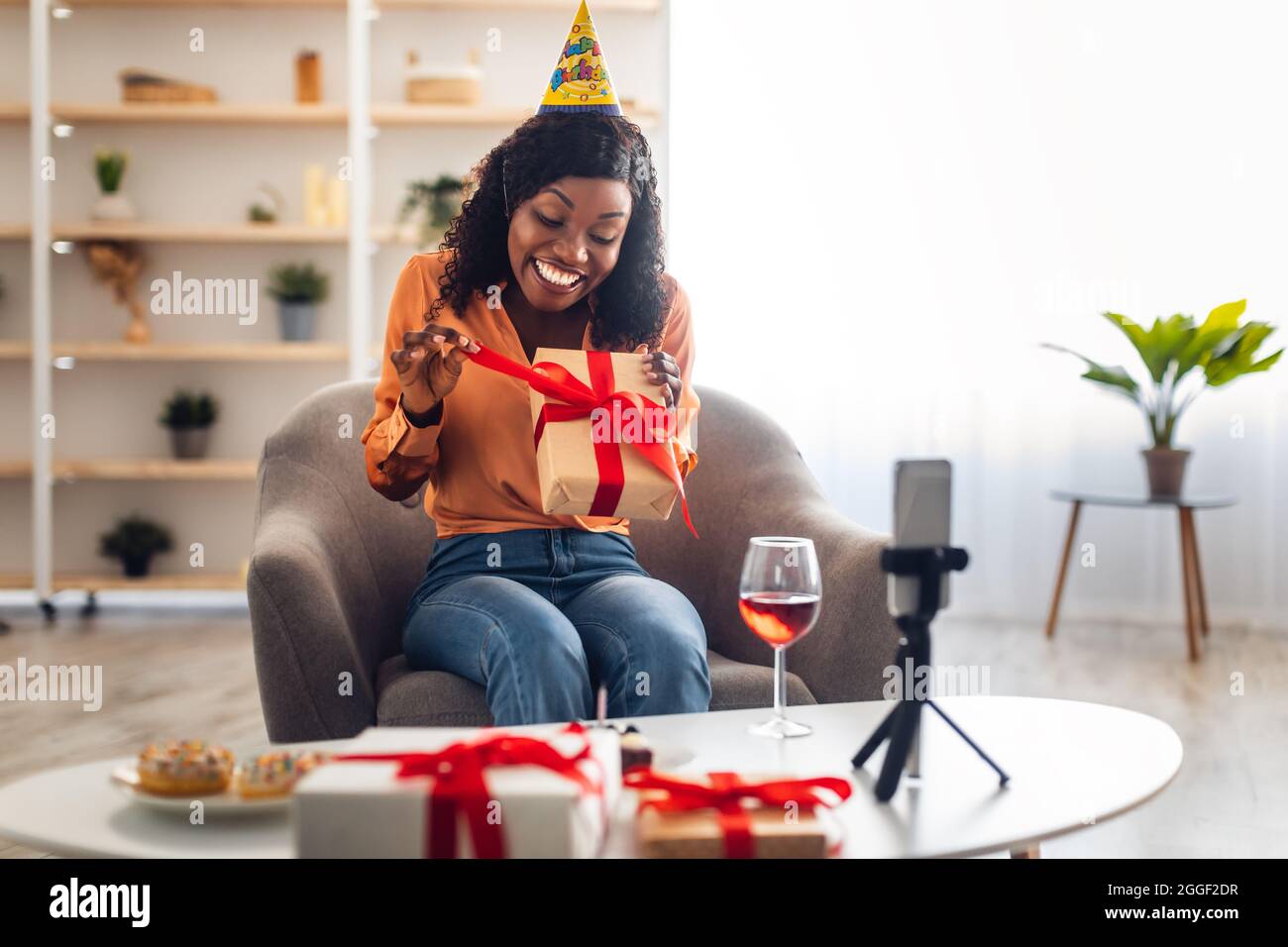 Donna nera sorridente che apre il regalo di compleanno di fronte allo smartphone Webcam che ha partito in linea via videochiamata seduta in soggiorno all'interno. Distanza ho Foto Stock