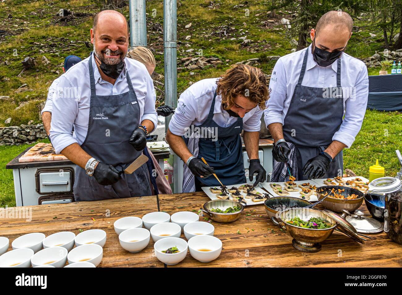 Tre chef del ristorante "miil" preparano piatti a un tavolo all'aperto a "Unplugged Taste" sulla Gompm-Alm in Alto Adige, Italia; piccole ciotole di salsa sono pronte per la degustazione. L'evento, che si tiene ogni anno l'ultima domenica di agosto, vede chef rinomati preparare i loro piatti su stufe di legno storiche. Foto Stock