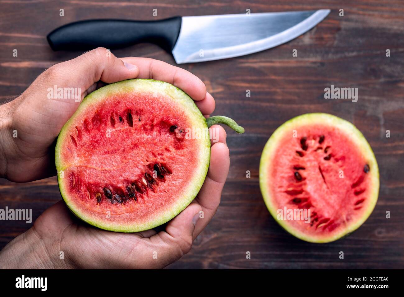 Metà del piccolo cocomero nelle mani degli uomini. Un altro mezzo di cocomero e coltello sul tavolo. Foto Stock