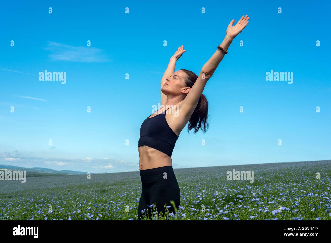 Donna sportiva con le braccia sollevate che si allungano in un campo con cielo blu. Foto Stock