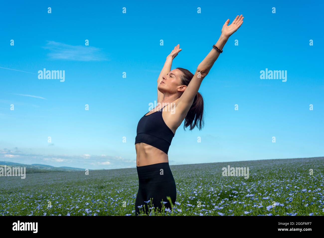 Donna sportiva con le braccia sollevate che si allungano in un campo con cielo blu. Foto Stock