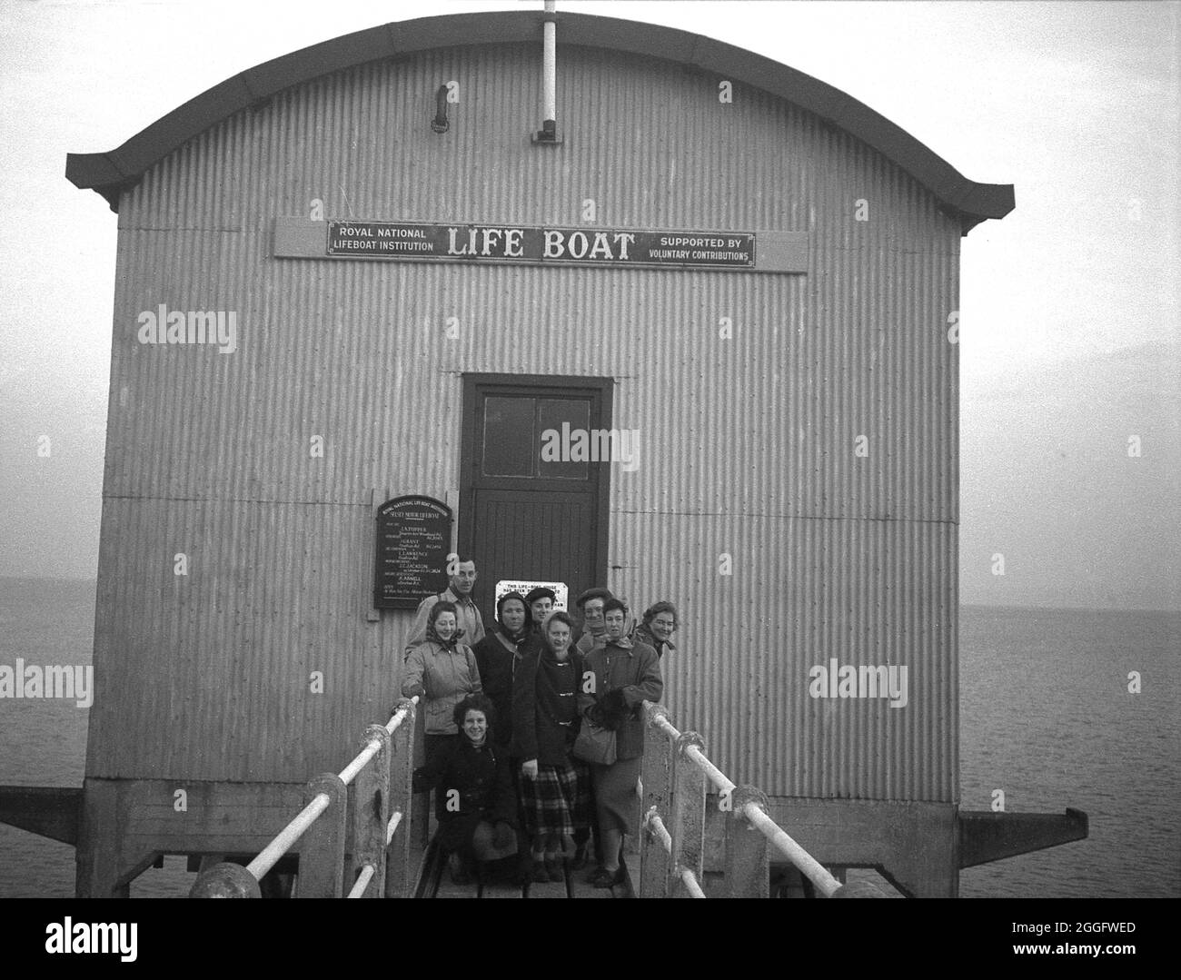 1950, storico, persone in piedi sulla passerella di legno di fronte alla vecchia stazione di scialuppa di salvataggio a Selsey, West Sussex, Inghilterra, Regno Unito. Un servizio di scialuppa di salvataggio è stato istituito a Selsey nel 1861. Nel 1927 per ospitare il nuovo motonave della stazione, la casa barca è stata costruita e questa struttura è visibile nella foto. Questa stazione di scivoli fu nuovamente ricostruita alla fine degli anni '50, ma fu abbassata nel 2017. Foto Stock