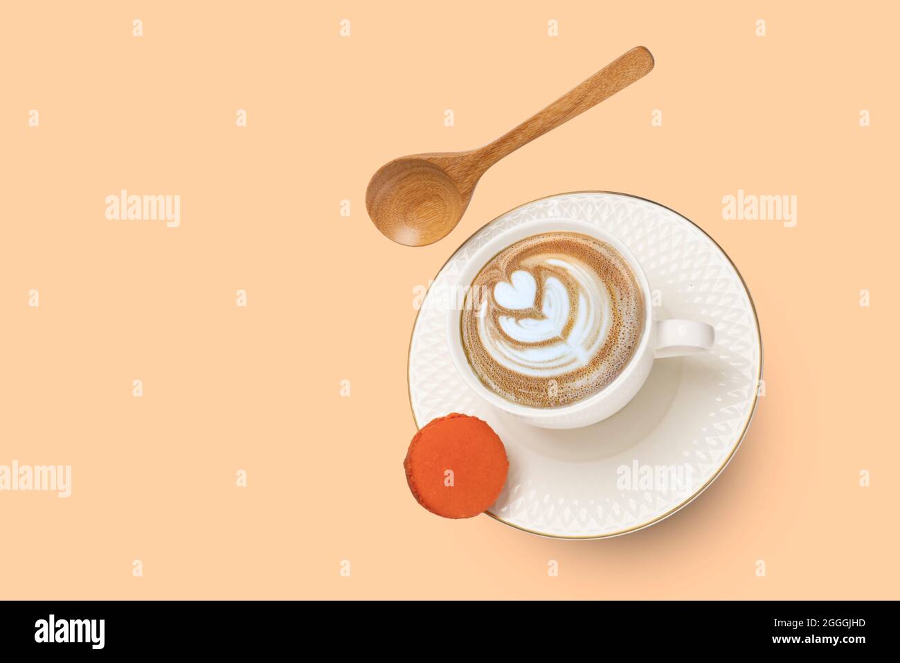 Una tazza di porcellana bianca con cappuccino e un piattino dorato con cucchiaio di legno e macarone arancio che si levita. Foto Stock