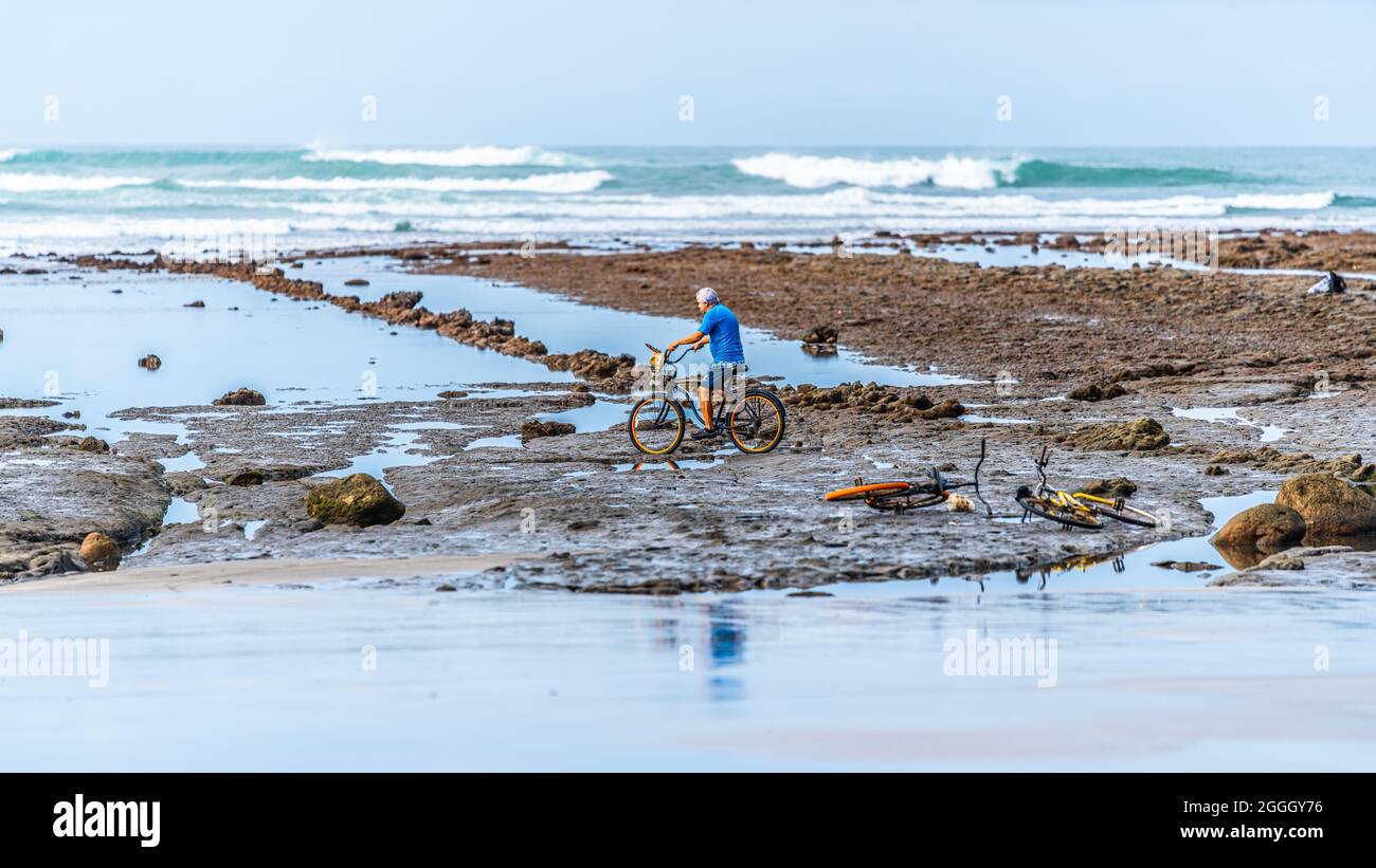 Un uomo del costa rica che indossa una t-shirt blu sta guidando una bici su una spiaggia rocciosa alla costa pacifica del Costa Rica. Durante la bassa marea. Foto Stock