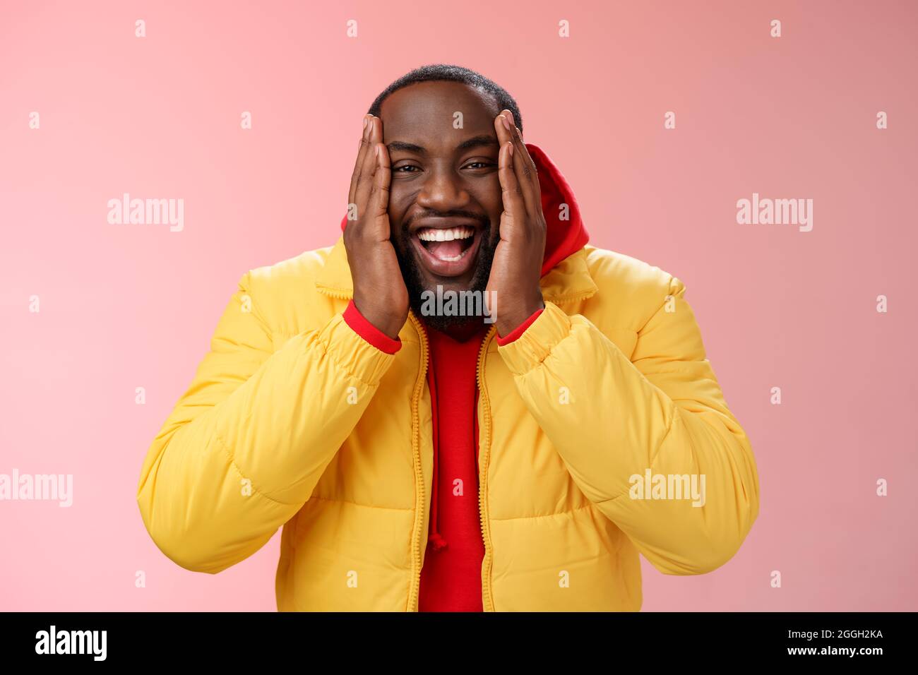 Felice carismatico giocoso afroamericano bearded ragazzo in giacca gialla red hoodie premere palme guance sorridenti amichevole ridendo felice scherzando non può Foto Stock