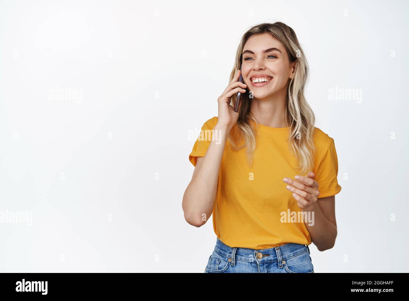 Concetto di tecnologia cellulare. Sorridente giovane donna che fa una telefonata, parla al cellulare con espressione del volto felice, indossa una maglietta gialla e dei jeans Foto Stock