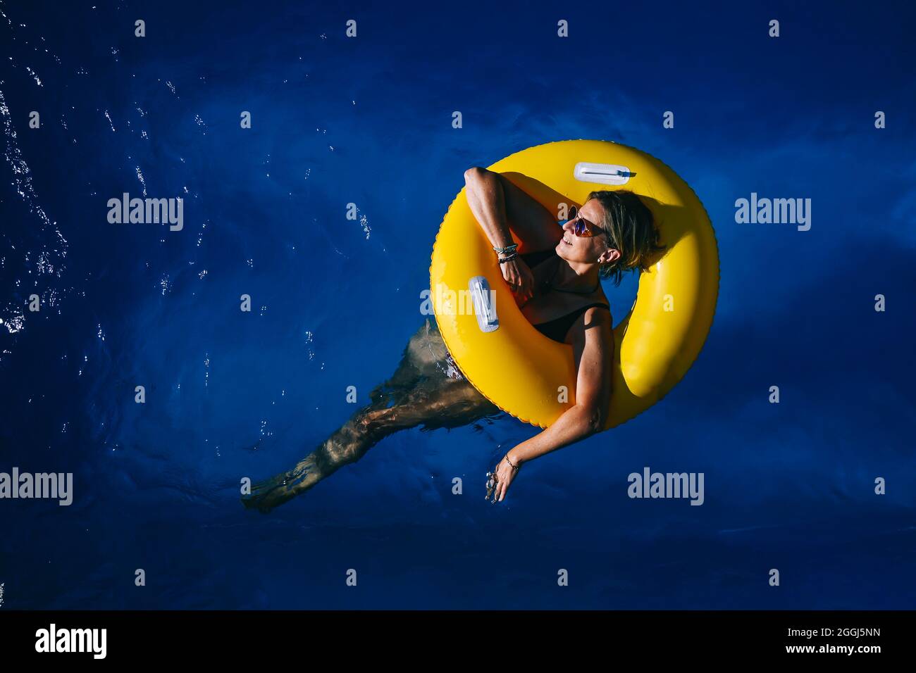 donna di 66 anni che si gode una giornata di sole in piscina con un galleggiante giallo Foto Stock