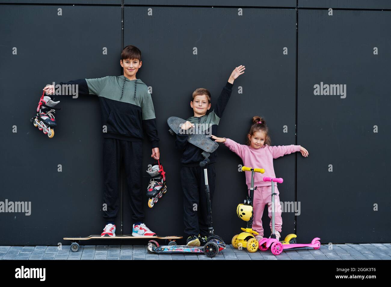 Tre bambini all'aperto contro il muro nero moderno. La famiglia sportiva può trascorrere del tempo libero all'aperto con scooter e pattini. Foto Stock