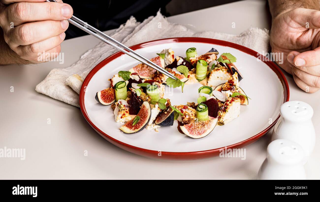 Striscione per alimenti. Le mani dello chef stanno preparando una gustosa insalata con fichi maturi, formaggio di capra, rotolo di cetrioli freschi, barbabietole e foglie di senape con t Foto Stock