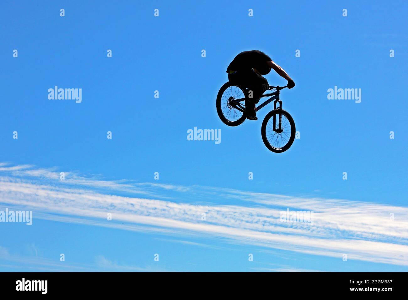 Silhouette di BMX o Mountain Biker in alto nell'aria durante uno spettacolo di acrobazie ambientato contro un cielo principalmente blu pallido con una banda di nuvole bianche Foto Stock