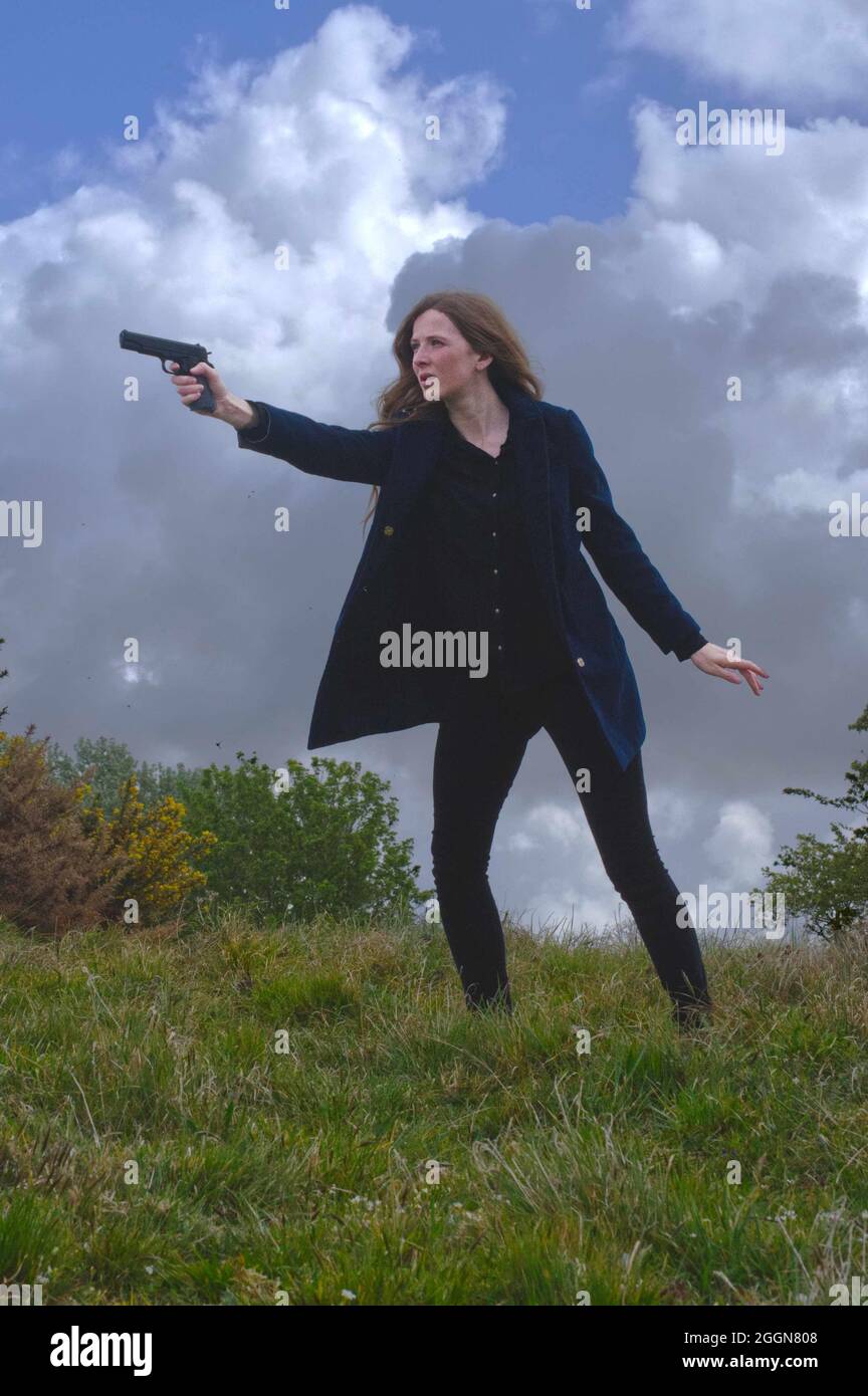 donna con pistola che indossa panni scuri su erba hillock con nuvole tempestose nel cielo (copertina libro) Foto Stock