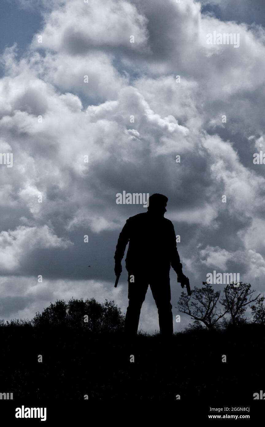 Silhouette di uomo in campagna in piedi contro un cielo nuvoloso, tenendo una pistola. (stile copertina libro) Foto Stock