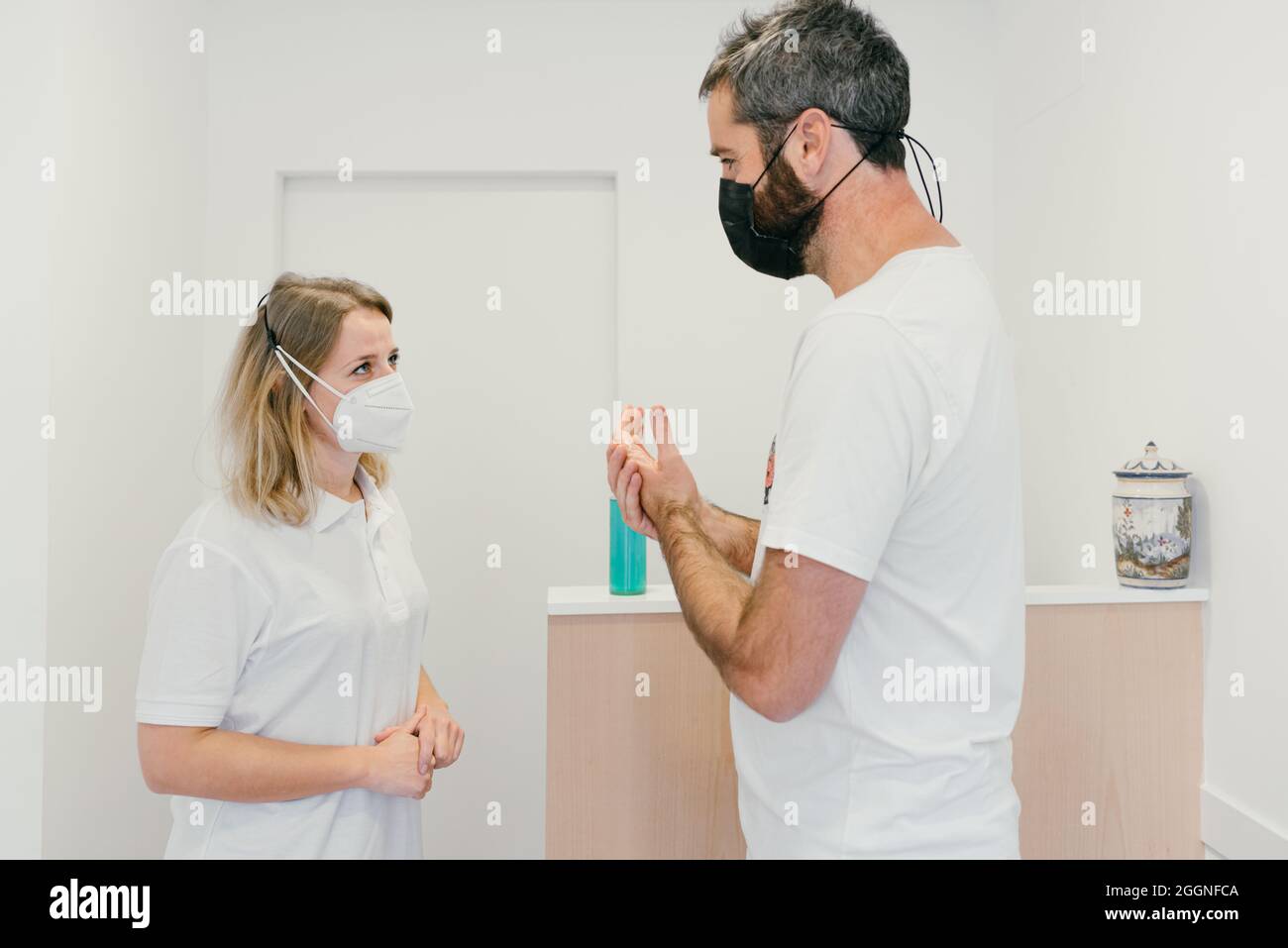 Gruppo di persone bianche che si parlano con la maschera e mantengono la distanza di sicurezza. L'uomo alto disinfetta le mani con gel idroalcolico. Foto Stock