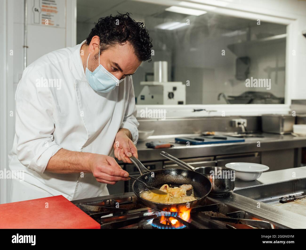 Il cuoco maschio si protegge con una maschera durante la sua giornata di lavoro nella cucina del ristorante. Lo chef afferra la padella con la mano sul fuoco. Foto Stock