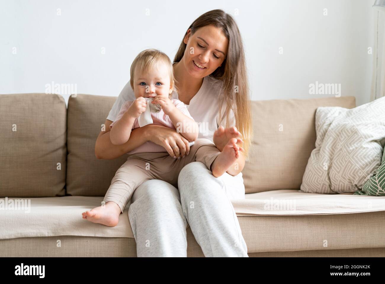 Madre sorridente dai capelli chiari seduta con il toddler biondo dagli occhi blu che mangia i calzini sul divano a casa Foto Stock