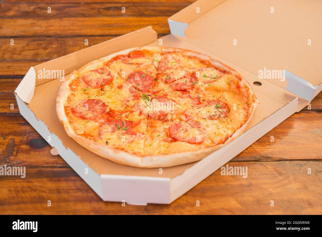 La pizza appena fatta con formaggio e salame in una confezione di cartone è un alimento veloce nocivo sullo sfondo di un tavolo di legno. Foto Stock