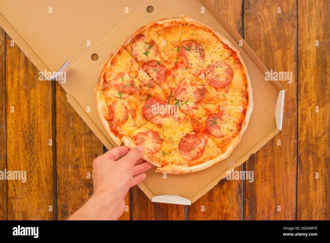 La mano del ragazzo prende una pizza in scatola di cartone di sullo sfondo di un tavolo di legno. Deliziosi fast food. Foto Stock