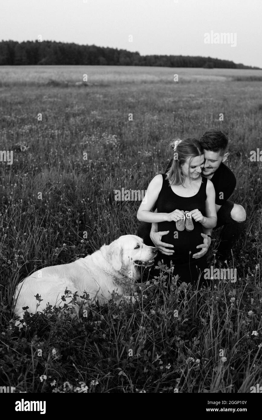Il giovane esprime tenerezza e cura alla moglie incinta, che tiene le scarpe dei bambini nelle sue mani, il loro cane più intelligente sta giacendo accanto a lei. B Foto Stock