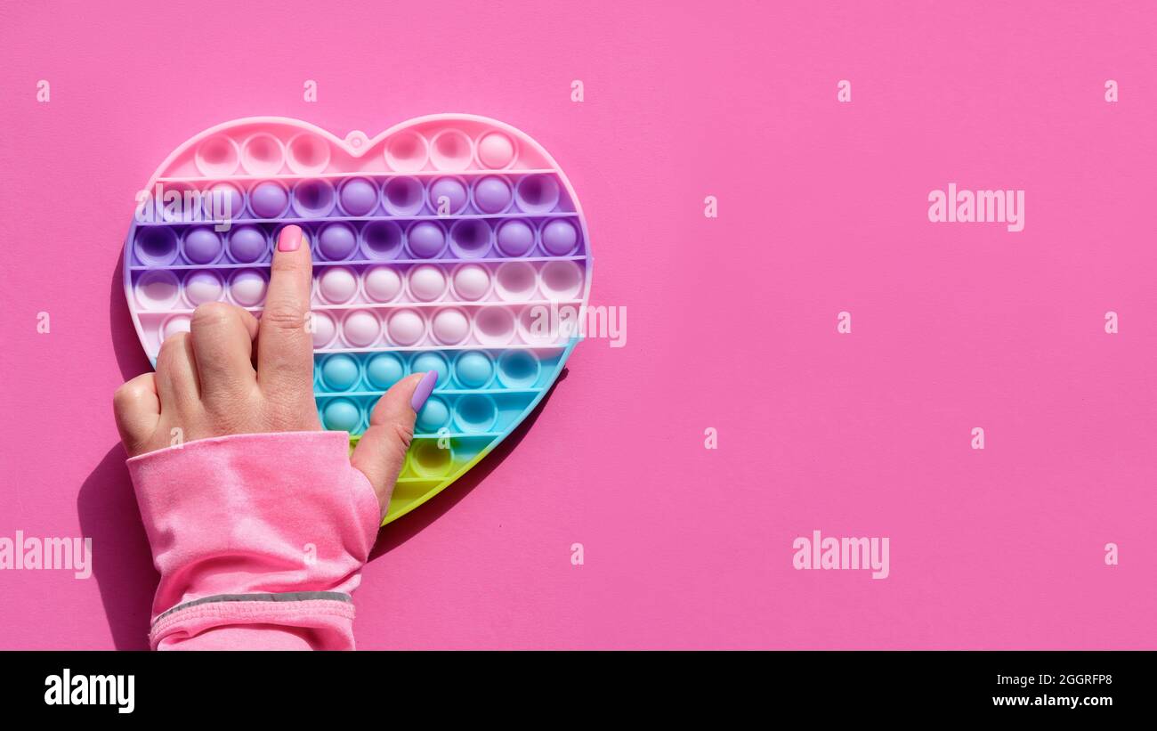 Cuore a forma di papa esso antistress giocattolo con mano femminile. Semplice fodget sensoriale a concavità nei colori di LGBT. Posare su carta rosa con spazio di copia Foto Stock