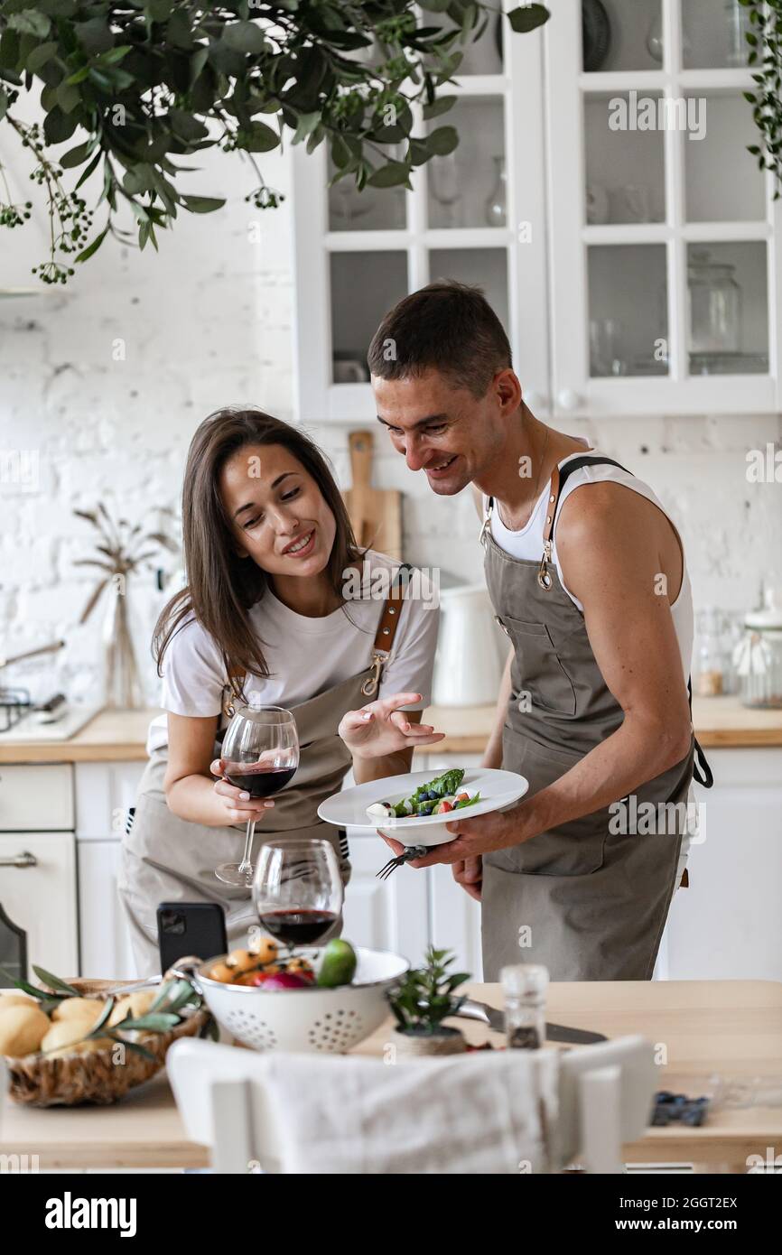 Coppia felice fare videochiamata con smartphone mentre si prepara la cena in cucina a casa allegra gente caucasica stanno agitando le mani, sorridendo Foto Stock