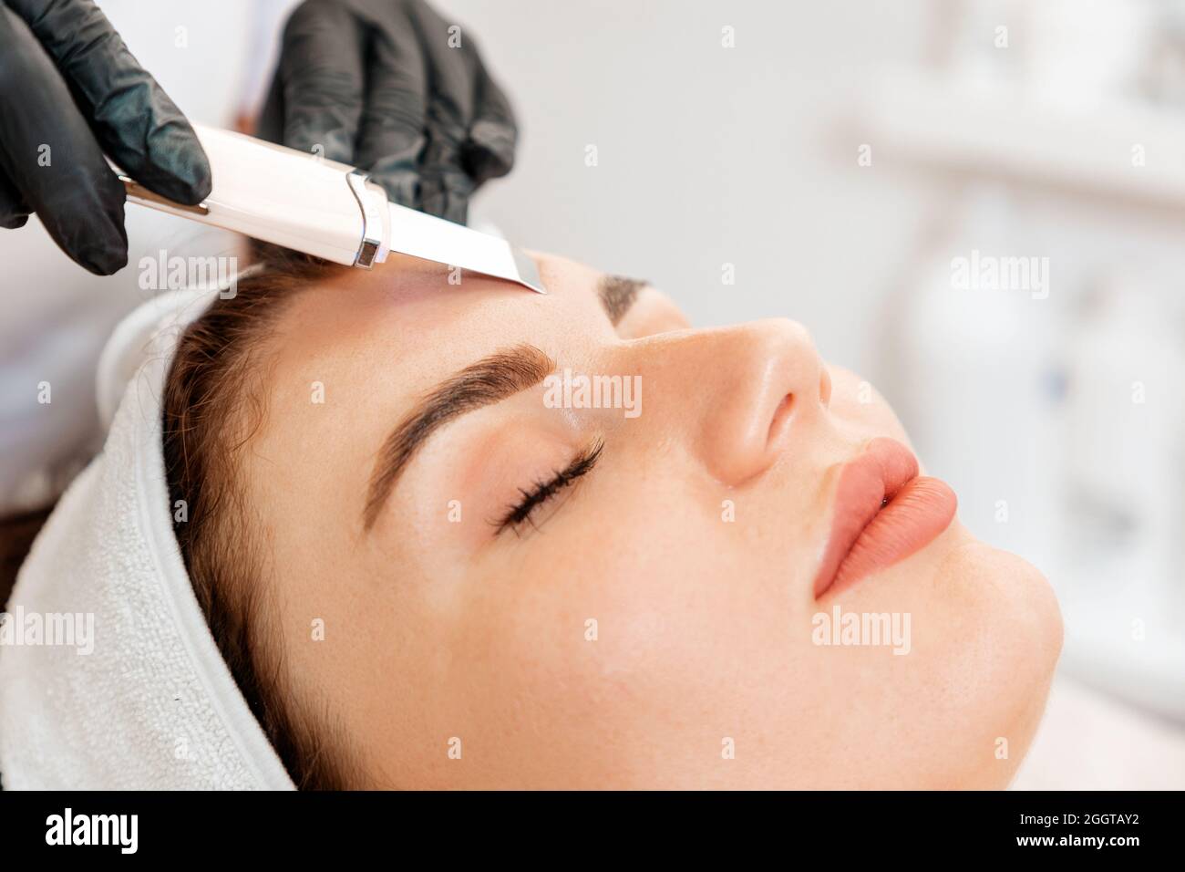 Procedura presso il salone di bellezza. Il cosmetologo pulisce la fronte del cliente utilizzando un dispositivo a ultrasuoni. Primo piano. Concetto di cosmetologia professionale. Foto Stock