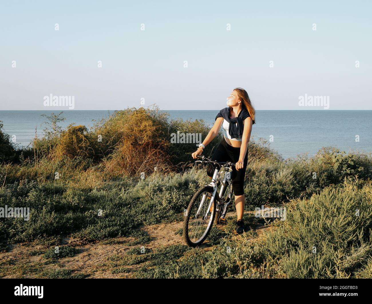 Giovane ragazza sportiva con una bici bianca sulla riva del mare in una mattinata estiva all'alba. Allenamento sportivo all'aperto. Immagine colorata. Foto Stock
