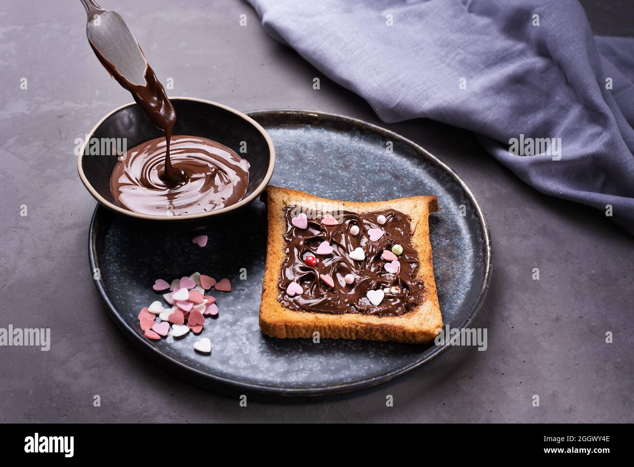 Pasta di cioccolato che gocciola da un coltello e toast con cacao spalmato con cuori multicolore su sfondo grigio, dolce dessert. Foto Stock