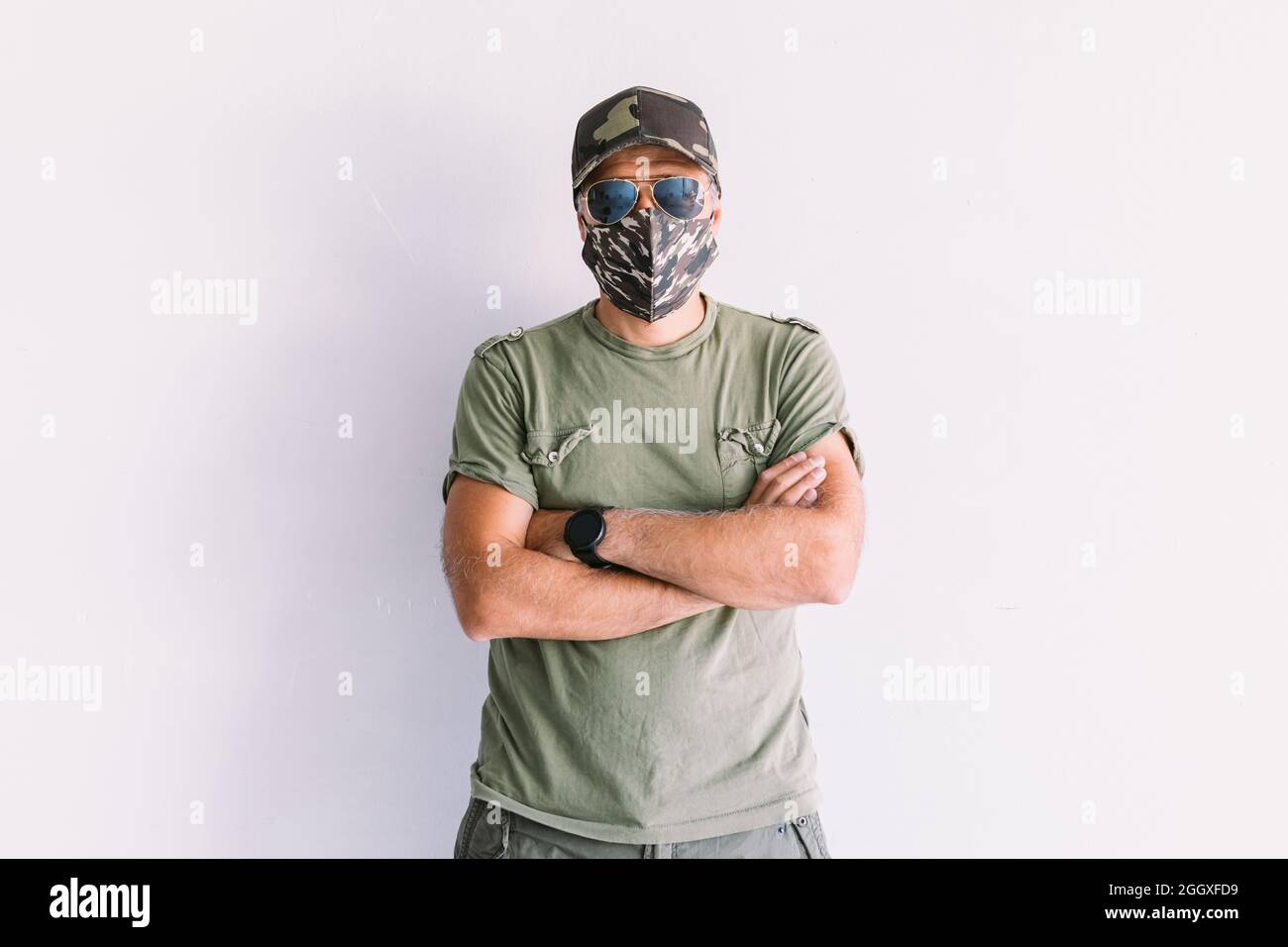 Uomo militare con cappuccio mimetico, occhiali da sole e maschera mimetica, a braccia incrociate, su un muro bianco Foto Stock