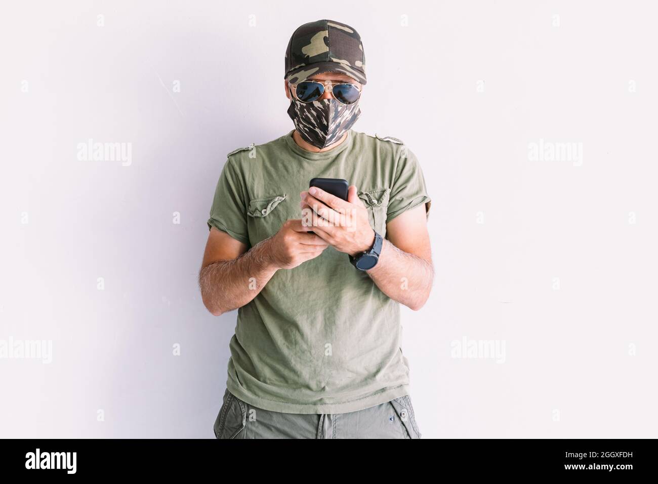Uomo militare con cappuccio mimetizzazione, occhiali da sole e maschera mimetizzazione, guardando il suo smartphone, su una parete bianca Foto Stock
