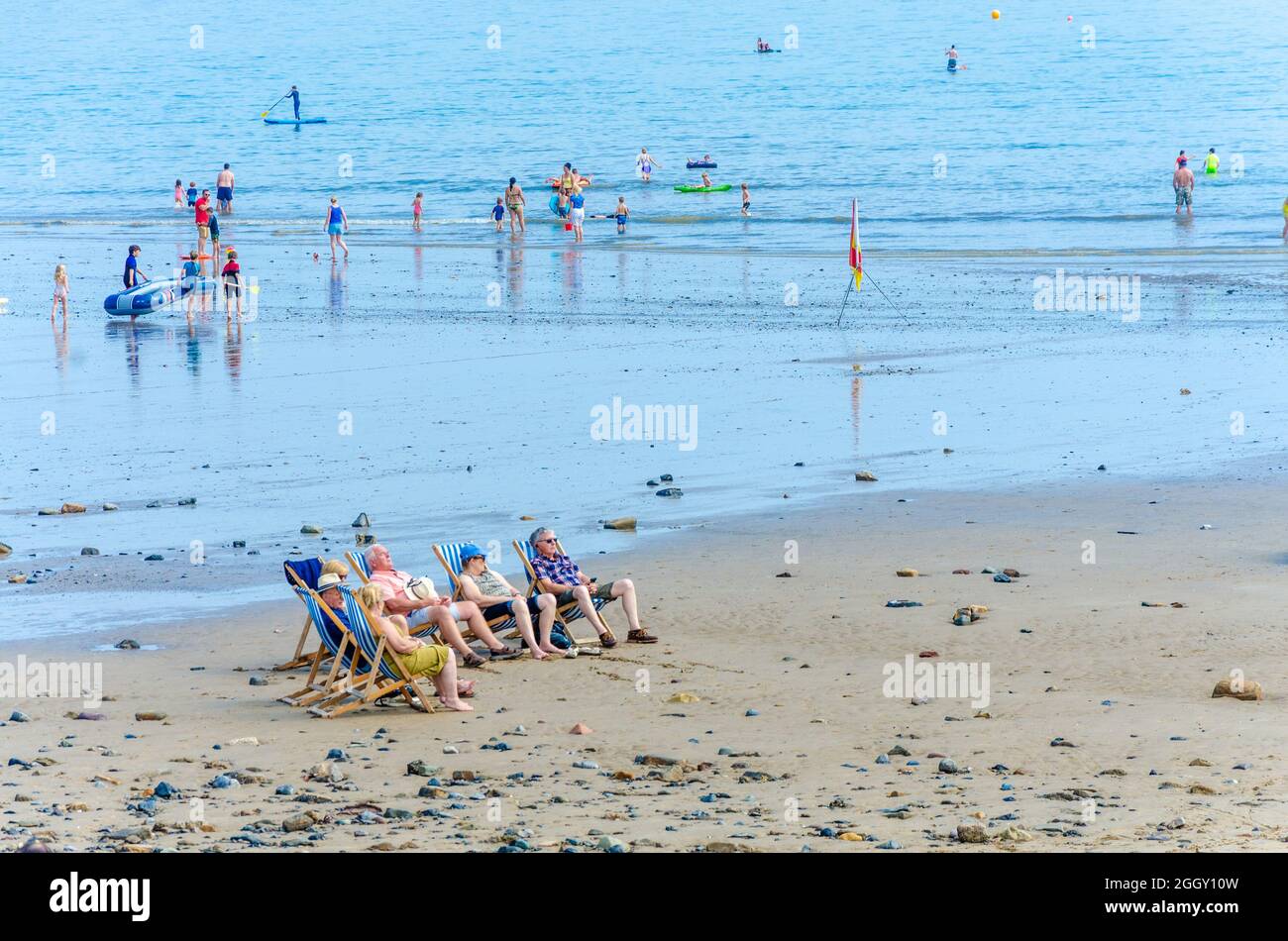 Un gruppo di amici si siede in sedie a sdraio sulla spiaggia di Saundersfoot a Pembrokeshire, galles, mentre le famiglie pagaiano in mare alle spalle. Foto Stock