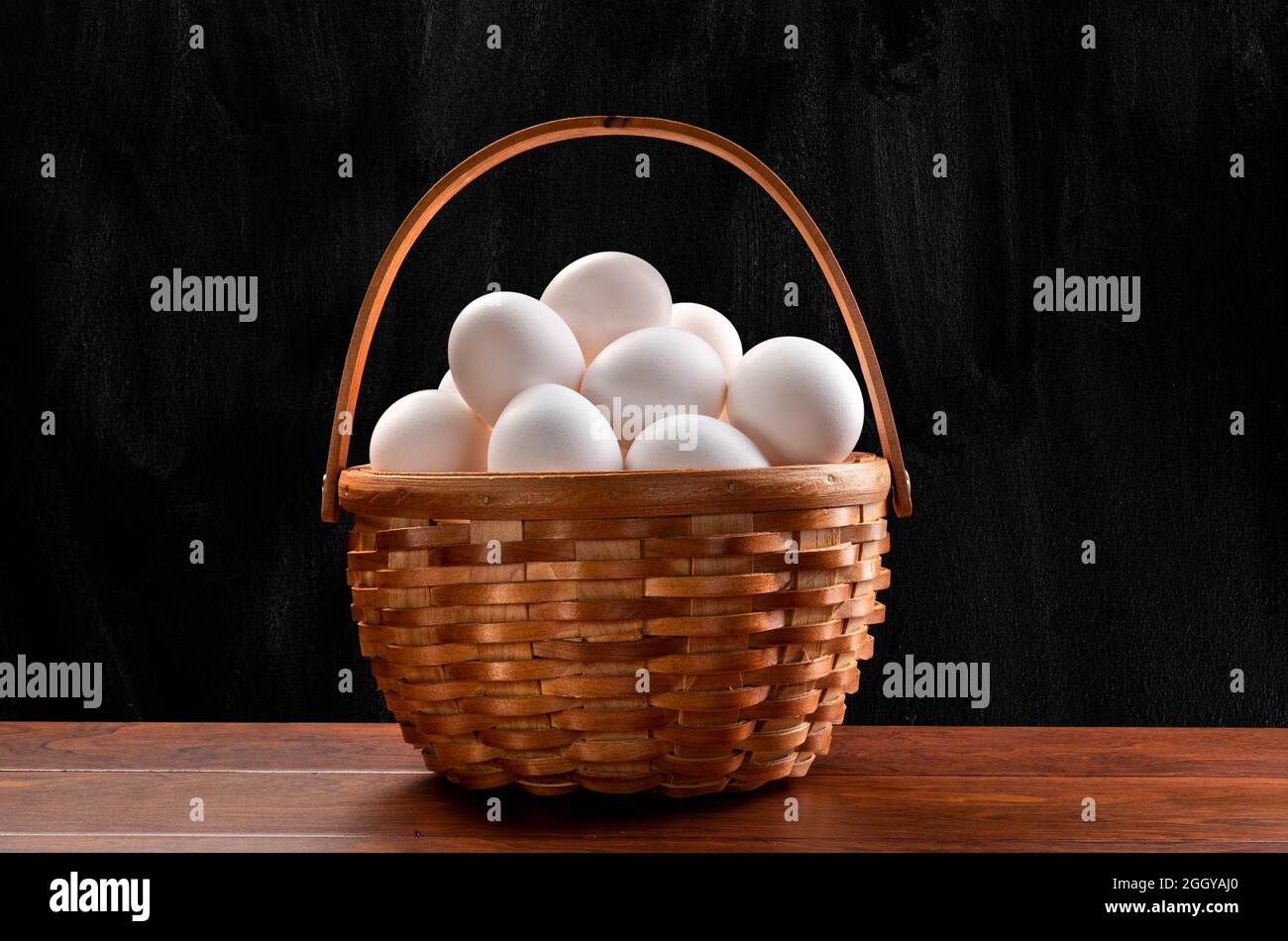 Un cestino di vimini di uova bianche appena raccolte sulla parte superiore di un tavolo di legno e incorniciato da un sottile sfondo nero, chiazzato. Foto Stock