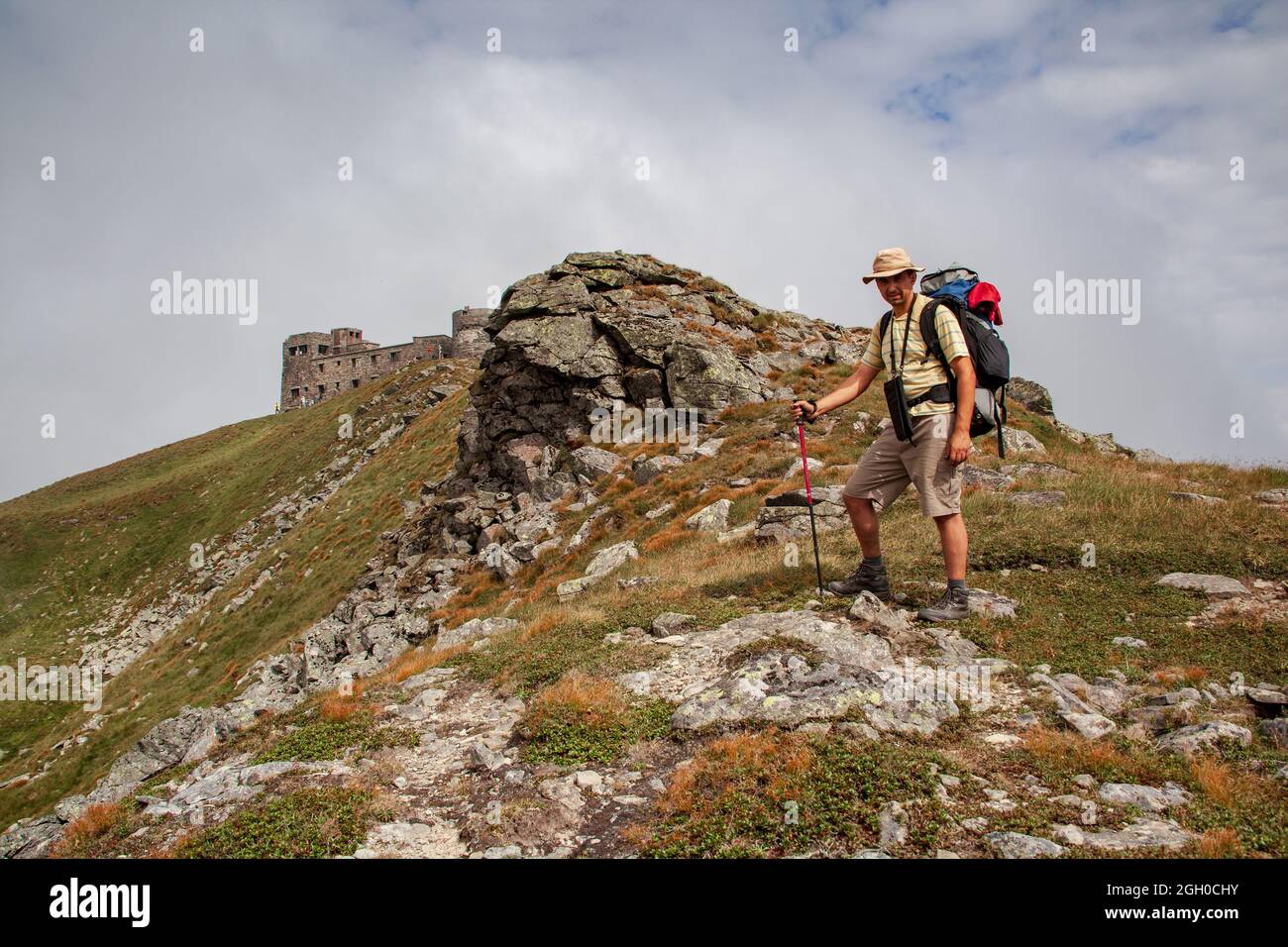 Uomo di mezza età con uno zaino sullo sfondo del paesaggio di montagna. Rovine dell'osservatorio astronomico chiamato elefante bianco sulla vetta - PIP Foto Stock