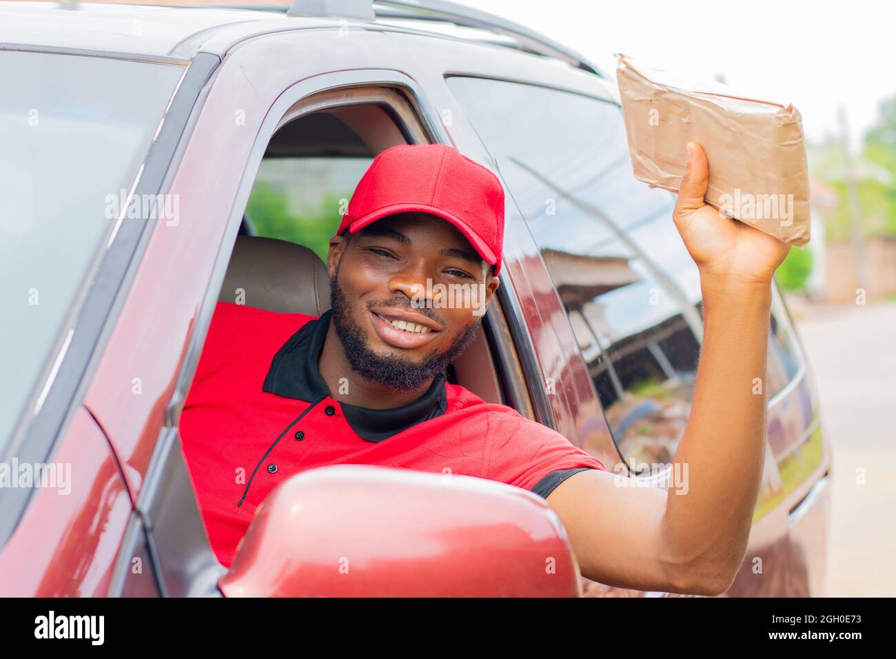 il lavoratore di consegna africano seduto in un'automobile che tiene un pacchetto e che agita a qualcuno per ottenere la loro attenzione Foto Stock