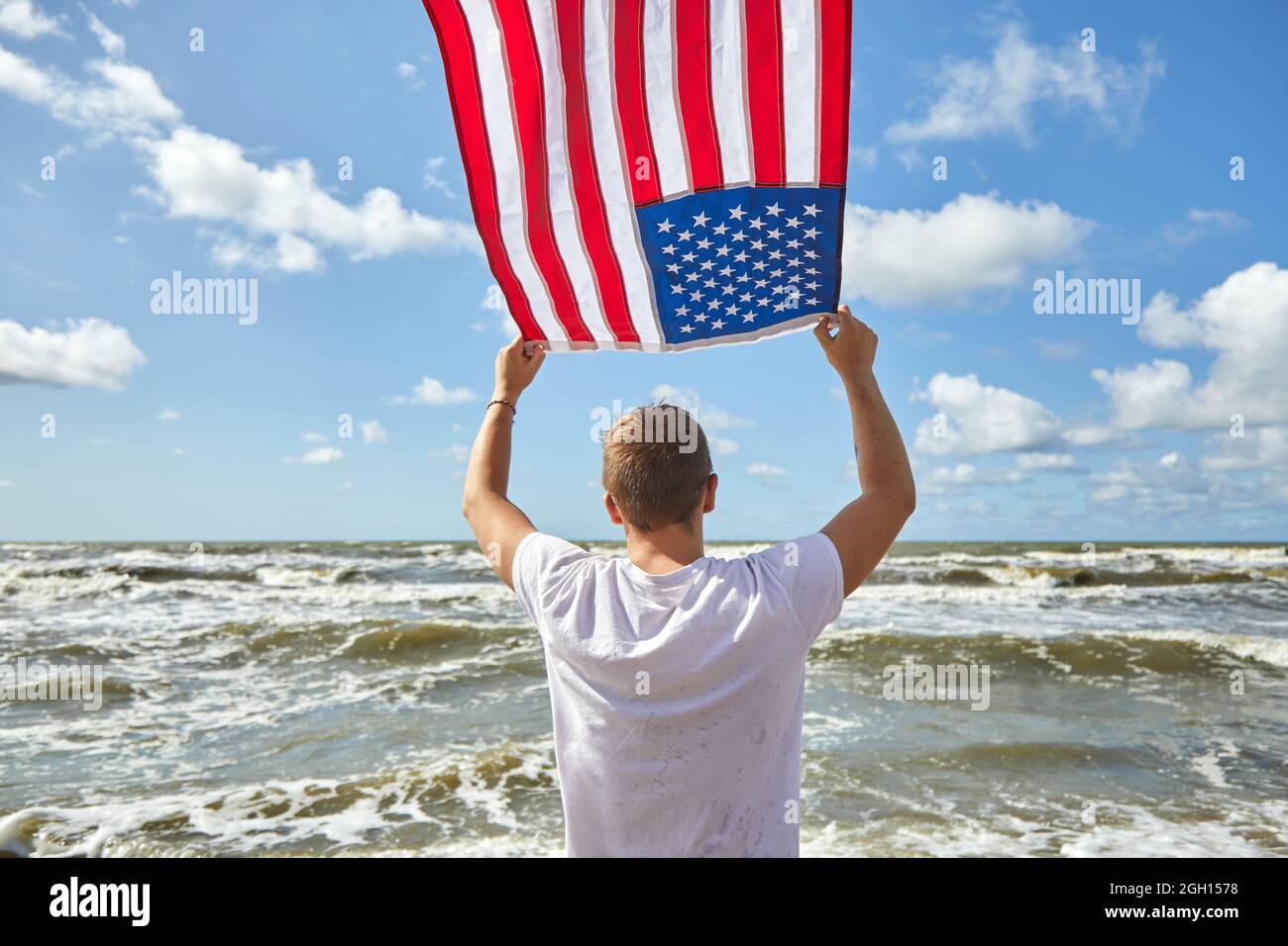 Uomo in abito bianco in piedi con bandiera americana ondeggiante nel vento sulla spiaggia. Foto Stock