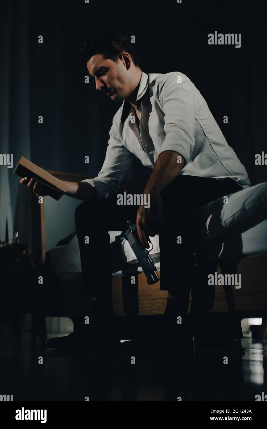 Un giovane uomo in abiti casual business seduto da solo in camera da letto tenendo una pistola e guardando la foto di famiglia in cornice sembrano andare ad uccidere se stesso. Foto Stock