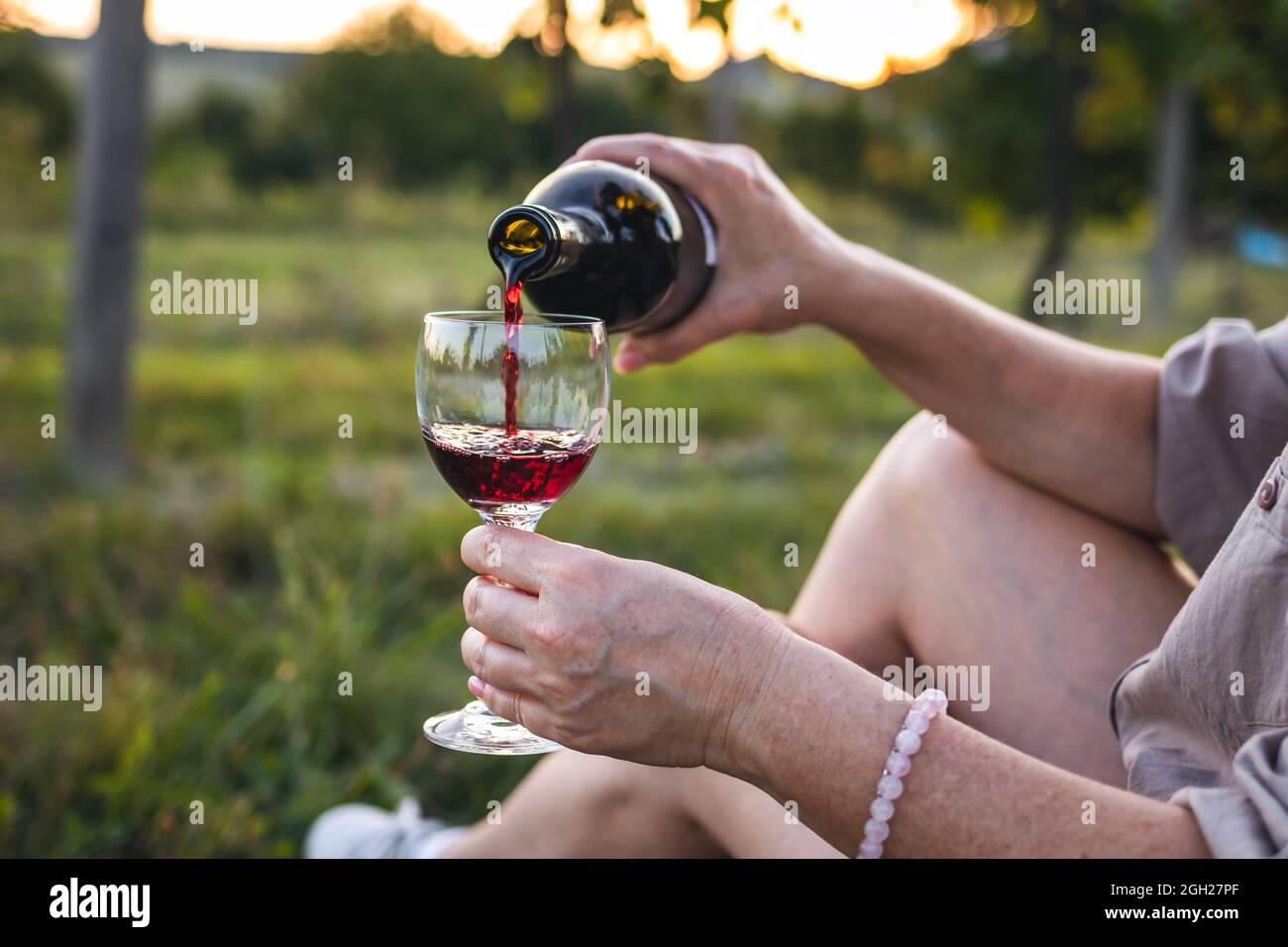 Donna seduta in vigna e versare il vino rosso in bicchiere da bere. Relax con bevande alcoliche durante il tramonto all'aperto Foto Stock