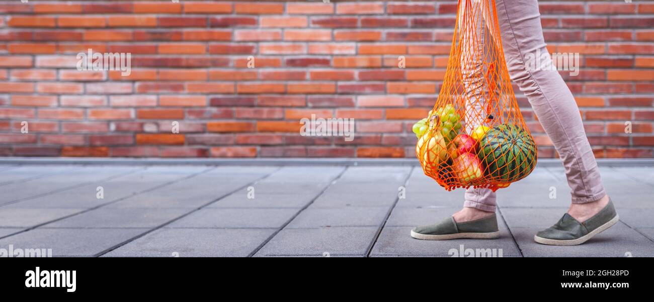 Donna che cammina con sacchetto riutilizzabile in rete pieno di frutta. Stile di vita sostenibile e concetto di zero sprechi. Sfondo muro di mattoni rossi. Foto panoramica Foto Stock