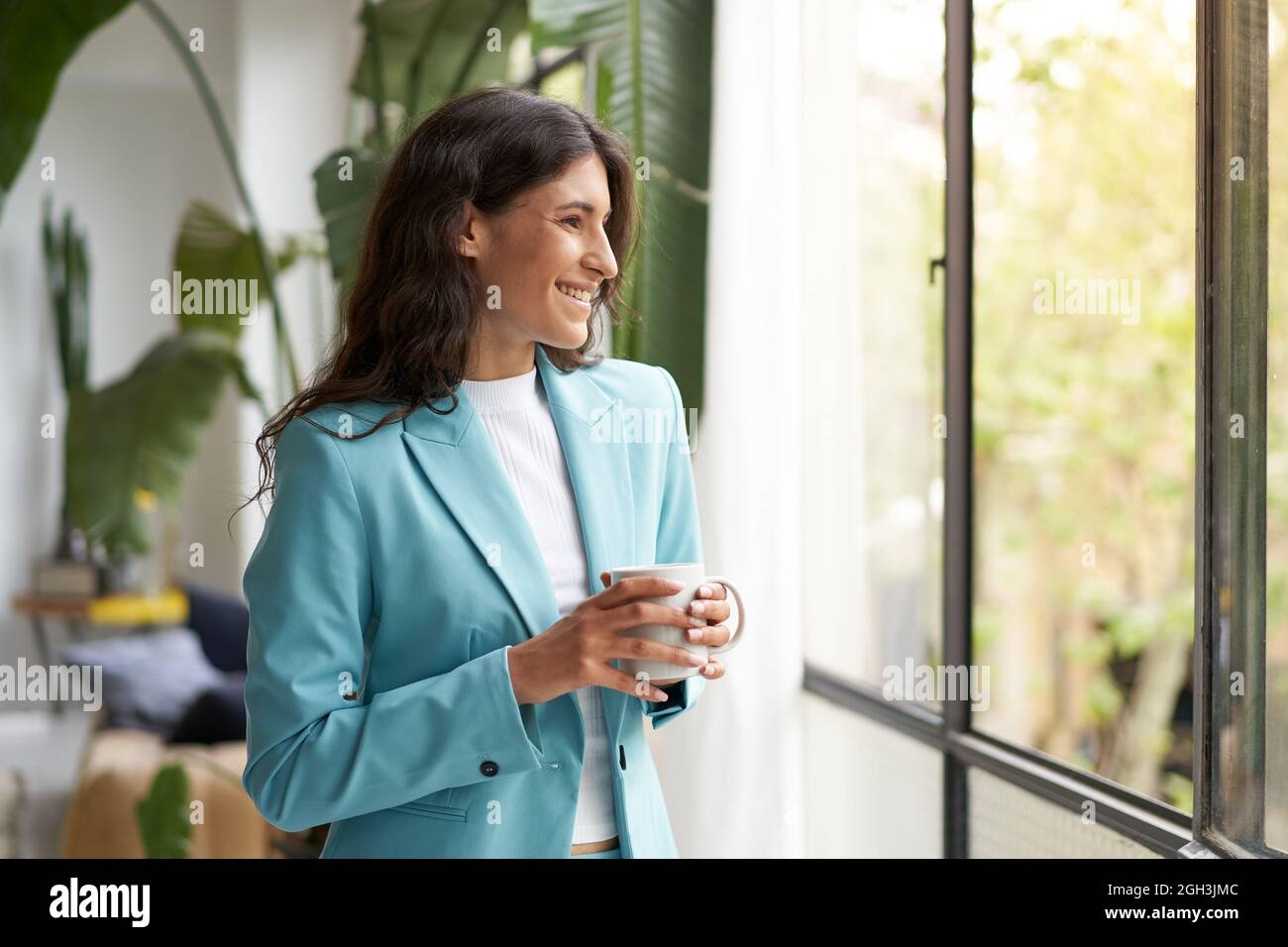 Giovane donna di affari sorridente soddisfatta di un lavoro ben fatto rilassarsi con il suo caffè o tè del mattino, guardando fuori la finestra. Bella latina donna Foto Stock