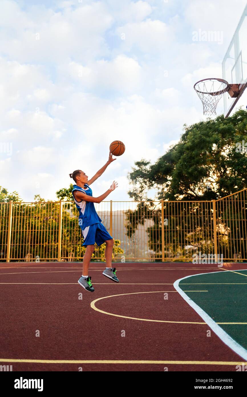 Basket. Un ragazzo di teenage in un abbigliamento sportivo blu salta e  getta una palla in una scarpa da basket. Campo sportivo sullo sfondo. Cielo  nuvoloso. Vista laterale. Co Foto stock -