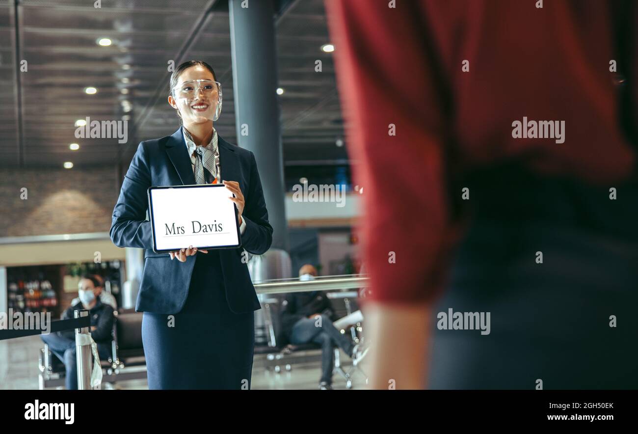 Donna che indossa uno schermo facciale in piedi al terminal dell'aeroporto, tenendo un cartello con il nome per ricevere un viaggiatore. Autista femminile con un cartello sul terminal degli arrivi. Foto Stock