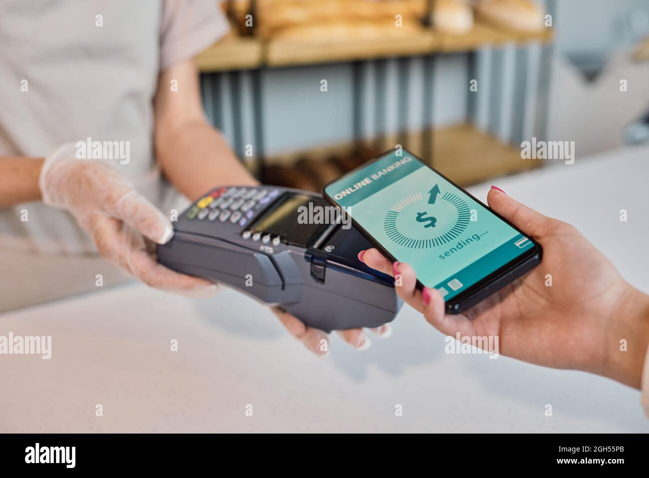 Primo piano di un dipendente di panetteria con guanto che detiene il terminale mentre riceve il pagamento dal cliente utilizzando il sistema di online banking al telefono Foto Stock