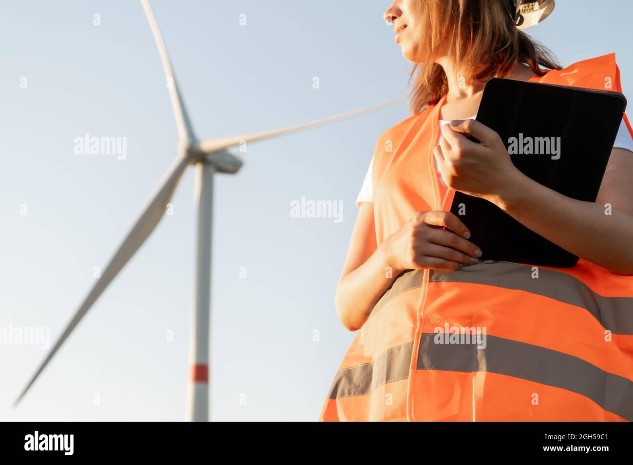 Donna nel gilet arancione e casco contiene documenti sulla manutenzione delle turbine eoliche sullo sfondo del mulino a vento alla luce del sole. Generazione di fonti rinnovabili Foto Stock