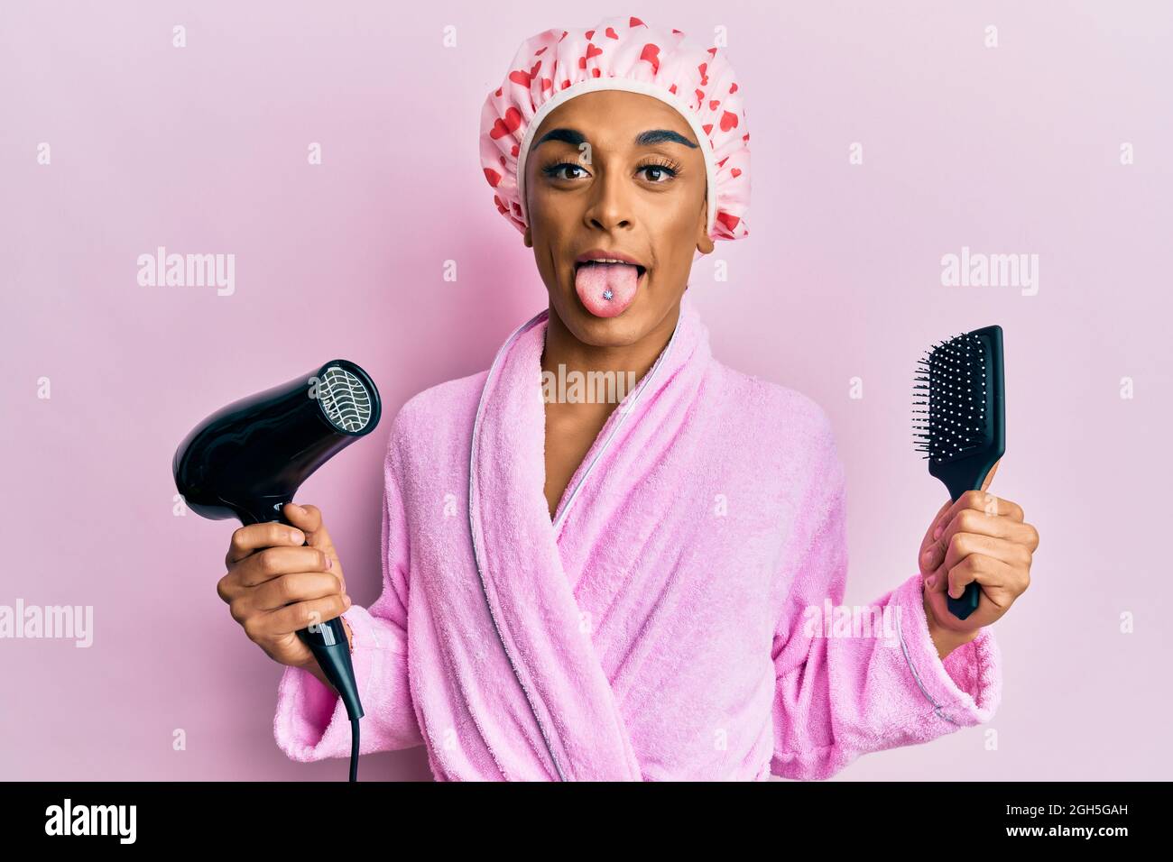 L'uomo ispanico che indossa il make up che indossa il cappuccio della  doccia che regge l'asciugacapelli e il pettine che attacca la lingua felice  con espressione divertente Foto stock - Alamy